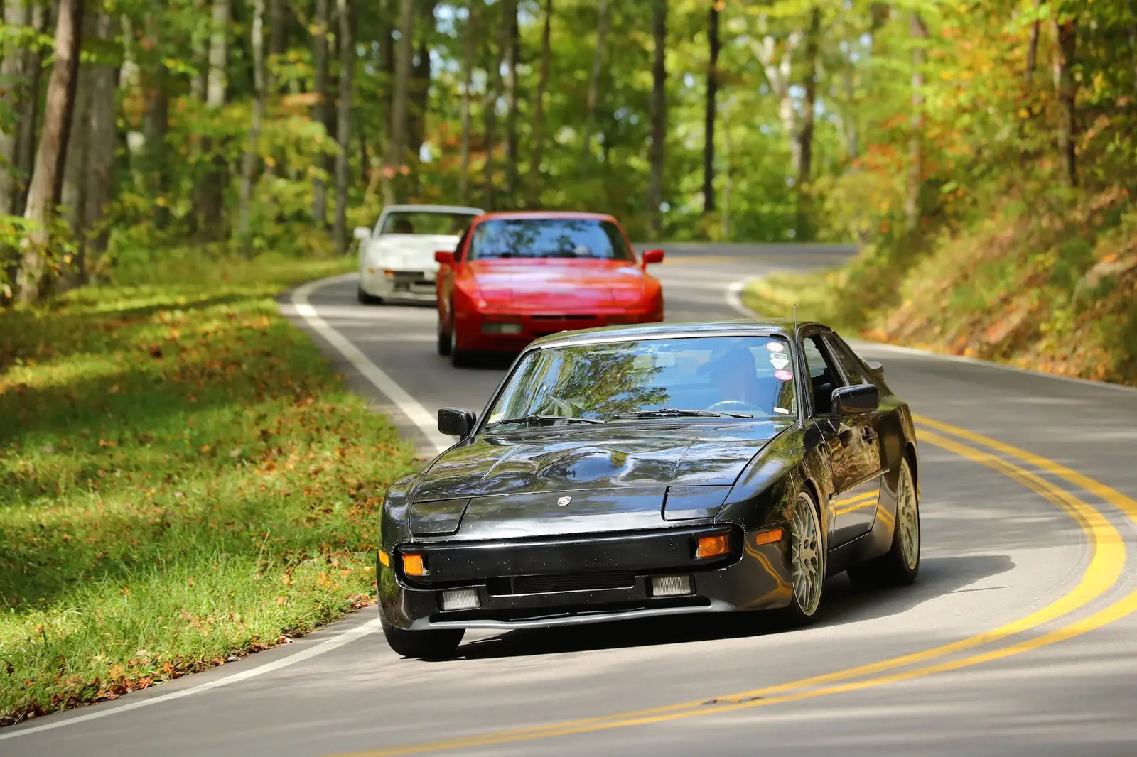 A black 944 leading a red 944 and a white one through a wooded curve