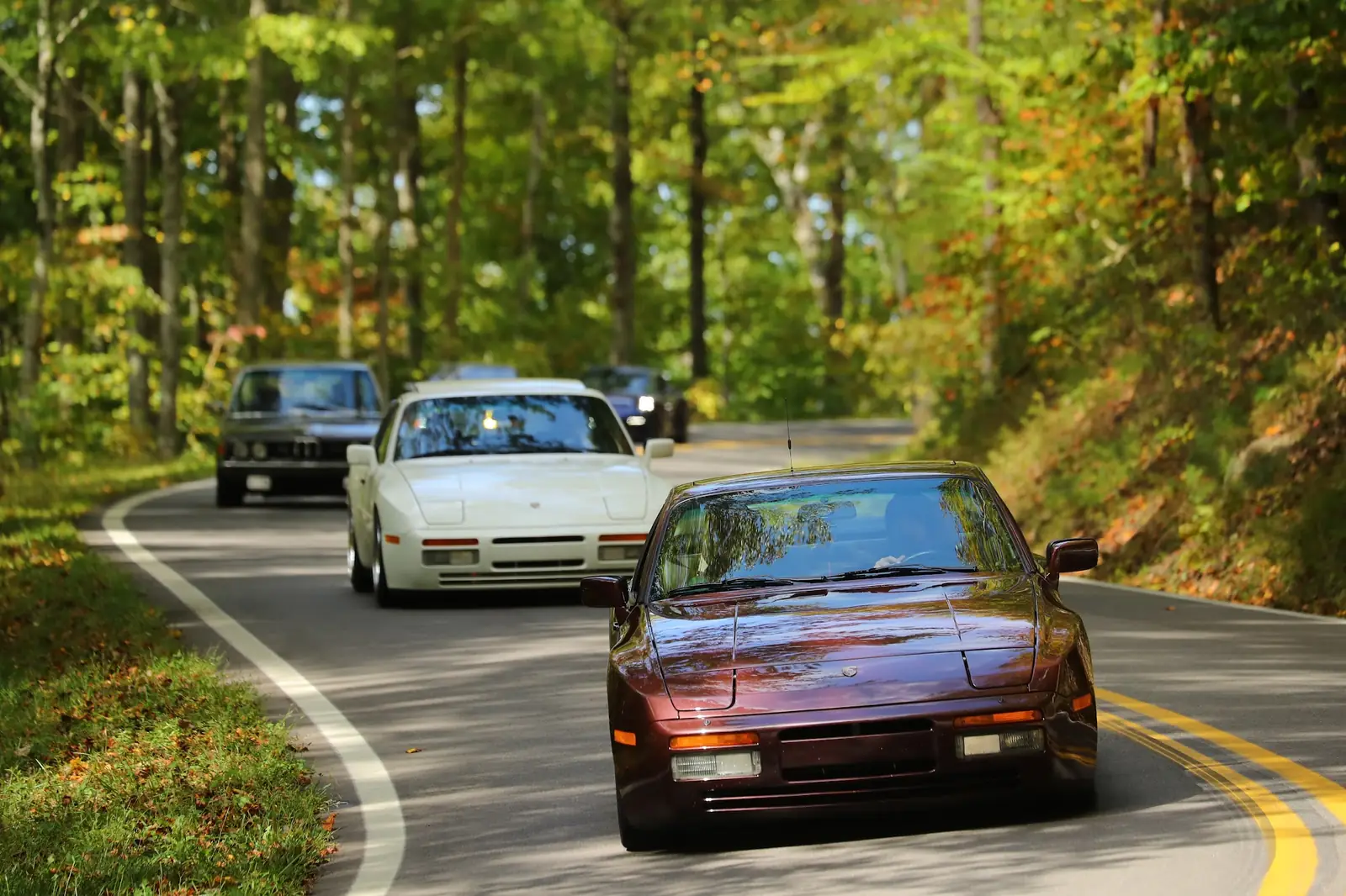 A maroon 944 leading a white 944 and a BMW sedan through the trees